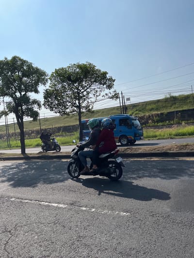 Two people are riding a motorcycle on a paved road in Pamotan, Indonesia, under a clear blue sky. The motorcycle is dark colored and the riders are wearing helmets. One rider is in front, wearing a green helmet, and the other is behind, wearing a blue helmet and a red jacket. A blue bus is visible driving on the road behind them, and to the right of the road, there is a grassy embankment with train tracks. Trees are on the left side of the road. The scene captures a typical moment of transportation in the Indonesian city.