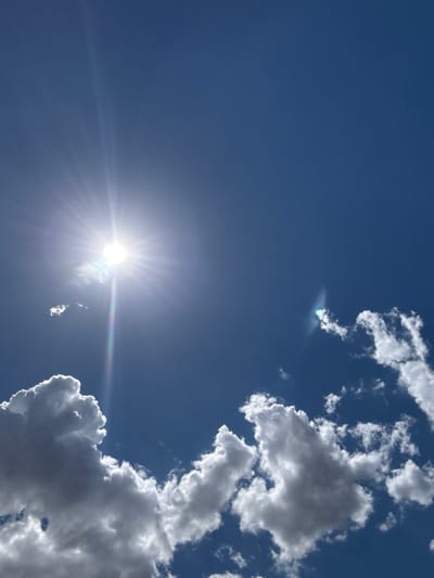 A bright sun radiates light and lens flare across a clear blue sky. A few fluffy white and gray clouds are scattered throughout the sky, with larger formations in the lower left and right quadrants. No people, structures, or landforms are visible. The location is not specified within the city of Бурсоль, Russia.