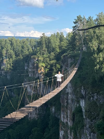 A person is walking across a suspended wooden bridge, which spans a deep gorge. The bridge is constructed with a series of wooden planks forming the walkway, supported by ropes and cables. The person, wearing a white top and dark pants, is positioned midway across the bridge, facing away from the camera and looking towards the right.
The scene is set in a natural environment with lush green trees covering the steep slopes of the gorge. The rocky cliffs are visible on the sides of the gorge, partially covered by vegetation. Above the trees, a bright blue sky with scattered white clouds indicates fair weather. The sunlight casts shadows on the bridge, suggesting it is daytime, likely late morning or afternoon. The overall impression is one of adventure and connection with nature, with the person seemingly enjoying the experience of crossing the bridge over the expansive landscape.
The location context is Kul'metovo, Russia, which hints at a potentially remote or mountainous area known for such natural attractions. No visible text is present in the image.