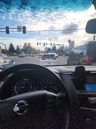 The image depicts an urban intersection in Fife, Washington, observed from the driver's seat of a Nissan vehicle. The vehicle is stationary, indicated by multiple red traffic lights visible ahead. The sky is partly cloudy with blue patches. In the foreground, the Nissan steering wheel, dashboard, instrument cluster (showing a lit green turn signal indicator), and a central console with a screen and phone mount are visible. Directly in front, a white Honda CRV with a visible Washington license plate is stopped in traffic. To the right of the CRV, a dark-colored passenger van is also stationary. On the right side of the intersection, a "76" gas station with a fuel price sign (partially showing "3.69") is prominent. On the left, a white monument with a partially visible "CITY OF FIFE" sign is present, along with street signs for "Pacific Hwy E" and "54th Ave E". Trees with some autumn foliage are visible in the background, and overhead power lines and traffic light gantries cross the scene. No individuals are clearly discernible.