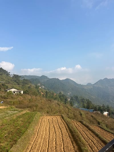 The image depicts a rural, mountainous landscape in Minh Tân Commune, Vietnam, under a clear blue sky with scattered white cumulus clouds. The foreground features agricultural terraces, with a cultivated plot on the left showing green growth, and a prominent central plot of freshly tilled, light brown soil with distinct parallel furrows.
On the left midground, a light-colored, single-story house with a dark roof is situated on a vegetated slope. Further up the left mountain, smaller, indistinct structures are visible. A lone individual, dressed in blue, is bent over in the green terraced field on the far left, engaged in an indeterminate activity.
The midground and background are dominated by multiple layers of densely tree-covered green mountains, stretching across the horizon. On a lower slope towards the right, a structure with a blue roof is partially visible, with a small plume of white smoke or haze rising nearby. Other distant, small structures are sparsely dotted across the mountainous terrain. A dark metal railing is visible in the bottom right corner, indicating the vantage point of the photographer. The overall scene suggests a clear day in a rural, agricultural mountain environment.
