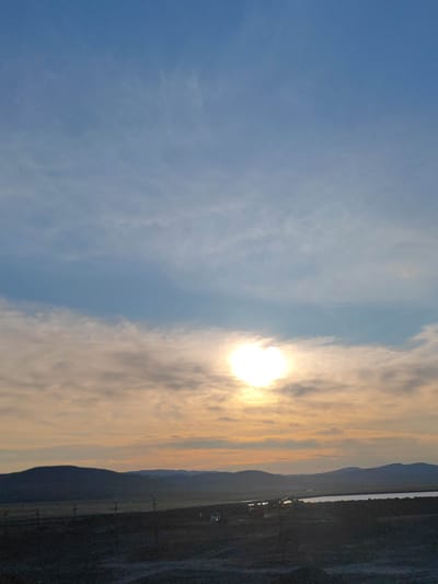 This is a vertical, full-frame photograph capturing a sunset or sunrise over a hilly landscape. The sky is a gradient of blue at the top, transitioning to a lighter blue and then to soft oranges and yellows near the horizon, where thin clouds are illuminated by the sun. The sun itself is a bright, diffused orb positioned slightly to the left of the center in the middle of the frame, partially obscured by clouds.

Below the sky, a dark, silhouetted landscape unfolds. Rolling hills form the horizon line, with several distinct peaks. In the mid-ground, there appears to be a body of water, reflecting the sky's light. Scattered across the foreground and mid-ground are several indistinct objects. To the left, in the foreground, there are what appear to be several upright crosses. Further into the mid-ground, there are some dark structures that could be buildings or industrial equipment, with some lights visible. On the right side of the mid-ground, a white vehicle, possibly a van or truck, is visible. The overall impression is one of a vast, open, and somewhat desolate environment.

The image is a landscape scene, likely taken outdoors in a rural or semi-rural area, given the natural topography and the presence of what appear to be memorial crosses. While the prompt mentions Mayskiy, Russia, there are no specific visual cues within the image itself to confirm this location. The scene is devoid of any explicit human activity or interaction.