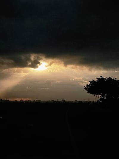 The image captures a dramatic sky over a dark, silhouetted landscape in Keude Ulim, Indonesia. Thick, ominous dark clouds dominate the upper portion of the frame, with a sliver of bright, golden sunlight breaking through the center, casting visible rays downwards. The sun appears partially obscured by the clouds, creating a powerful contrast between light and shadow.
Below the sky, the horizon is a dark silhouette of the land, featuring a line of trees and possibly buildings or structures. The foreground is almost entirely black, with the detail obscured by darkness, suggesting it is either very early morning or late evening. A solitary tree with a full canopy is visible in silhouette on the right side of the image, adding depth to the scene. The overall impression is one of a powerful natural phenomenon, with the human presence in the landscape minimal and largely unseen due to the low light conditions.
