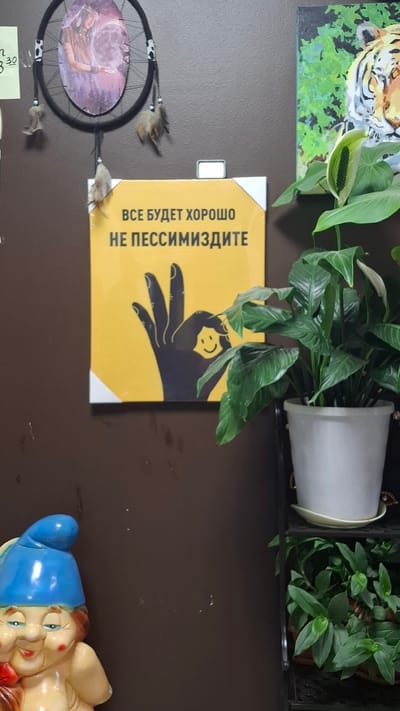 The image shows a wall with several decorative items. In the upper left, a dreamcatcher with feathers hangs on the wall. Below it, a yellow sign with black text reads "ВСЕ БУДЕТ ХОРОШО НЕ ПЕССИМИЗДИТЕ" which translates to "Everything will be fine, don't be pessimistic." The sign also features a black silhouette of a hand with a smiley face on it. To the right of the sign, a painting of a tiger's face is visible. In the foreground, a potted plant with large green leaves is placed on a shelf unit. In the lower left corner, a garden gnome figurine with a blue hat and red cheeks is positioned. The wall appears to be a dark brown color. The image was taken in Niukzha, Russia.