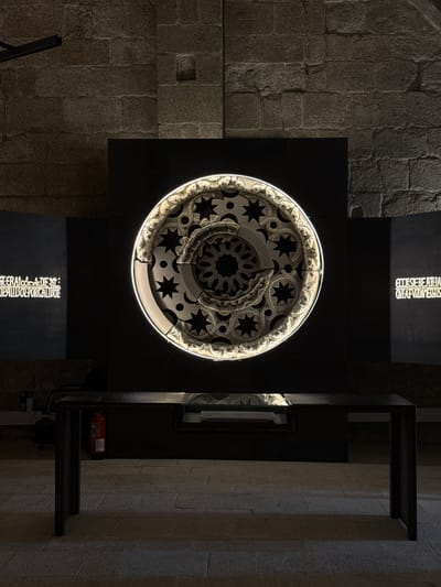 The image shows an interior view of a building, likely a historical site or museum in Santiago de Compostela, Spain. The main subject is a large, circular, illuminated art installation with intricate geometric and star patterns, made of what appears to be carved stone or plaster. The installation is mounted on a dark, rectangular structure against a textured stone wall. To the right, a dark, multi-level display shelf unit is visible, with a red fire extinguisher placed on one of the shelves.
The lighting is dim, suggesting an indoor setting with artificial illumination. The stone walls, one appearing rough-hewn and the other smoother and more regular, provide a sense of age and permanence to the location. The artwork's complex design and illumination create a focal point, hinting at an exhibition or display.
Visible text is present on two illuminated panels. One panel at the top, oriented horizontally, reads "CITES EST BEATA ESTER..." followed by more text that is partially obscured or difficult to discern clearly. Another panel at the bottom, oriented vertically, displays text that appears to be in Latin, reading "SEMPITERNAM COGNOSCITE DEI: / EIUS PRINCIPALIUS ET PORRA ILLUDE". The presence of this text suggests an informational or explanatory aspect to the display, possibly related to religious or historical themes given the location.
The overall mood is one of quiet contemplation and historical reverence, amplified by the dim lighting and the juxtaposition of ancient stonework with modern artistic presentation. The time of day is not evident, but the indoor setting and artificial lighting are the primary environmental cues.