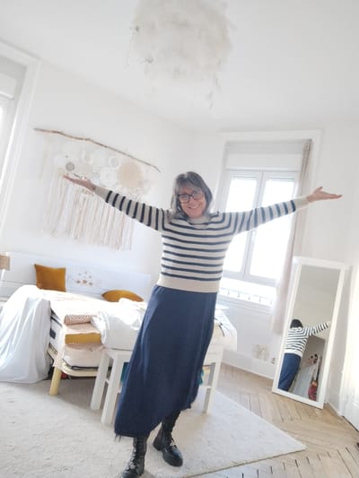 An interior scene depicts an adult female posing in what appears to be a bedroom or private residence in Montélimar, France. The female, centered in the frame, is wearing a long-sleeved cream and navy blue striped sweater, a long navy blue skirt, and black combat boots. She has dark, shoulder-length hair with visible gray and wears black-framed glasses. Her arms are extended horizontally outwards, and she is smiling directly at the camera. The room features white walls and a light-colored wooden floor, partially covered by a rectangular light-colored rug. Two large windows with white frames are visible, one on the left and one on the right, admitting bright natural light. A white bed frame with a white headboard and light bedding, including two mustard-yellow throw pillows, is positioned on the left. Above the bed, a decorative wall hanging consisting of a natural tree branch from which white and beige macrame or crochet elements and fabric strips suspend is mounted. A white, cloud-like feathered light fixture hangs from the ceiling in the upper center. On the right, a full-length, white-framed mirror stands, reflecting the woman and parts of the room. A small white stool or table is located in the foreground, near the bed.