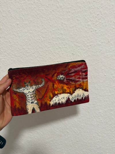 A hand holds a rectangular, fabric-covered object against a textured, white wall. The object's surface features a vibrant, abstract painting in shades of red, yellow, orange, and white. The artwork depicts a stylized, humanoid figure with red, flame-like hair or a headdress descending from the top left of the object. Below this figure, a quadrupedal animal with black and white stripes, resembling a tiger or zebra, is shown in a dynamic, mid-leap or running pose, its head turned to the left. The background is a mix of fiery reds and oranges with patches of white and green, possibly suggesting a chaotic or energetic scene. The fabric appears to be a tapestry or printed cloth, with visible texture and stitching along the edges. The bottom edge of the object shows signs of wear and slight fraying. The lighting suggests an indoor setting, possibly daytime, as indicated by the even illumination and lack of harsh shadows. The overall impression is of an artistic creation, possibly a pouch or a wallet, displayed against a plain backdrop. No text is visible on the object or the wall.
