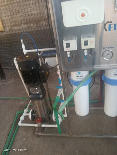 A water filtration system features a pump with a black motor and silver cylindrical body. It is connected to a network of white and green pipes, valves, and fittings. The system also includes two white filter housings with blue caps, mounted on a stainless steel panel. The panel has pressure gauges and electronic components. A date of "2026/02/11 08:21" is visible in the bottom left corner, likely from a camera's timestamp.