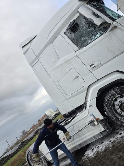 A single male individual is depicted washing the driver's side of a white Scania XG semi-truck cab. The truck's exterior is covered in white cleaning foam. The man, possessing a beard and dressed in a dark blue jacket, blue jeans, a black beanie, and light-colored work gloves, holds a long-handled brush and is actively scrubbing the lower section of the cab. The washing occurs on a wet, paved surface, exhibiting foam residue. In the background to the left, a metal fence borders a grassy area, beyond which a large array of solar panels is visible. Distant buildings are partially obscured under an overcast sky. This scene is located in Ostend, Belgium.