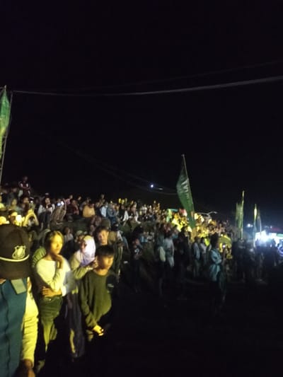 The image depicts a large outdoor gathering of people at night in Kecamatan Pujut, Indonesia. A dense crowd, composed primarily of young adults and some children, occupies an uneven landscape, possibly a hillside or natural incline, with many individuals standing or sitting.
The scene is illuminated by artificial lights, casting bright glows on sections of the crowd while leaving other areas in deep shadow, consistent with a nighttime event. The dark sky overhead shows subtle traces of power lines or cables. Several green flags are visible throughout the crowd, mounted on poles. One prominent flag clearly displays the text "TVRI," indicating an association with Televisi Republik Indonesia. Many attendees are looking in the same direction, suggesting a focal point such as a stage or screen is present but outside the frame. Some individuals are seen holding up their smartphones, possibly recording or photographing the event. In the left foreground, a person wearing a dark uniform, including a cap with a badge, is partially visible, potentially indicating security or event staff. The overall atmosphere suggests an engaged and attentive audience at a public event.