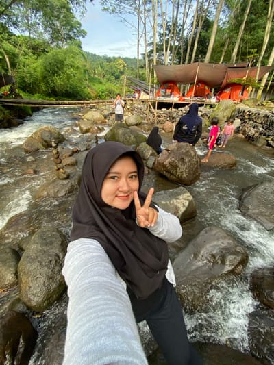 A young woman wearing a black hijab, a grey long-sleeve shirt, and black pants sits on rocks near a stream, flashing a peace sign at the camera. Behind her, a flowing stream cuts through a green, lush landscape. Some people are visible in the background. A small wooden bridge crosses the stream further back. Tents are visible in the background.