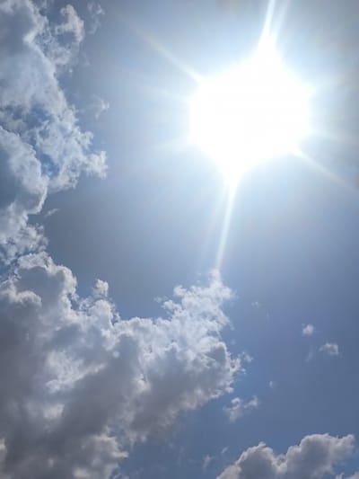 A photograph taken from a low angle, looking up at a bright, sun-drenched sky with scattered white and gray clouds. The sun is the dominant feature, positioned in the upper right quadrant of the image, emitting powerful rays that create a lens flare effect. The sky is a vibrant blue. The clouds are fluffy and appear to be moving, with some denser formations in the lower left and upper left of the frame. The overall impression is that of a clear, sunny day, possibly with some cumulus clouds forming. The image does not contain any identifiable landmarks that would specify a location within Alagyaz, Armenia.