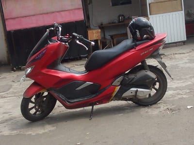 A red Honda PCX scooter is parked on a paved surface in front of a building. A black helmet with "PRO" written on it is resting on the seat of the scooter. The scooter has a license plate that is partially visible. The building appears to be a shop or a house, with a red and black facade. The environment suggests a street or a parking area in an urban or semi-urban setting. The time of day appears to be daytime, with natural light illuminating the scene. The location is identified as Megamendung, Indonesia.