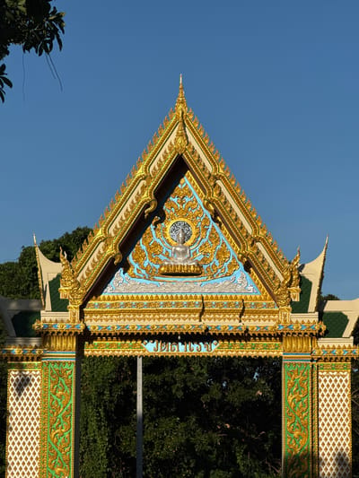 This image depicts the ornate entrance to a temple in Baan Tha Po, Thailand. The entrance features a steep, triangular roof adorned with intricate golden decorations and blue accents. At the apex of the roof, a silver Buddha image sits within a circular frame surrounded by golden floral patterns. The entrance is supported by two pillars, also richly decorated with gold and green ornamental designs. The background is a clear blue sky and lush green trees. The overall impression is one of elaborate craftsmanship and religious significance.