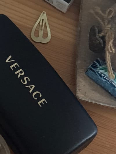 The image shows a dark blue case with the word "VERSACE" printed in gold lettering diagonally across its surface. To the left of the case, a gold-colored barrette with a teardrop shape is visible. The objects are placed on a wooden surface. The background is blurred, but appears to be a rough texture, possibly a box or cardboard, with a piece of twine and a small dark object. No people or specific locations in Enevo, Bulgaria are visible.