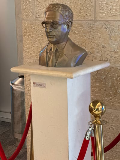 The image shows a bronze bust of a man wearing glasses and a suit, displayed on a white pedestal. A sticker with the text "TONY3D" is affixed to the pedestal. To the left of the pedestal is a silver trash can with a perforated surface. A red velvet rope barrier is visible in the foreground and to the right, with a brass stanchion topped by a golden sphere. The background consists of a stone wall. The location is identified as the Tzafria, Israel city square, a public area for events and gatherings.