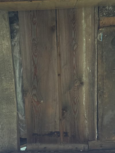 The image shows a close-up, overhead view of a weathered wooden structure in Ptichnik, Russia. The primary focus is a vertical, planked wooden surface, likely part of a wall or door. The wood exhibits prominent grain patterns, knots, and evidence of age and exposure, with some areas showing discoloration and what appears to be peeling paint or a weathered finish. To the left of the main wooden section, a portion of another wooden plank, lighter in color and rougher in texture, is visible. Behind the main wooden planks, a crumpled sheet of grey plastic sheeting is partially visible, suggesting a barrier or insulation. The background on the right shows a dark, textured surface, possibly brick or rough plaster, with some debris. The overall scene conveys a sense of disrepair and rural utilitarian construction. There are no people or animals depicted.