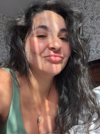 The image is a close-up, overhead shot of a young woman lying down, with her face angled towards the camera. She has dark, wavy hair that frames her face and cascades around her. Sunlight streams across her face, creating bright highlights and shadows, suggesting it is daytime. She is wearing a light green, ribbed tank top. A small, delicate tattoo of a sun and moon with a flower is visible on her upper chest. The background appears to be a bed with patterned bedding and a white sheet. The woman's expression is relaxed and serene, with her eyes partially closed and a gentle smile.