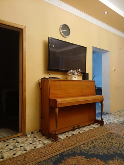 This is a full-shot, interior photograph taken from a low angle. The scene depicts a living room with a light yellow textured wall and a white ornate ceiling molding. On the wall, a flat-screen television is mounted above a wooden upright piano with a bench. A round clock with a black face and silver hands is visible on the wall to the left of the television. A bouquet of pink and white flowers in a white vase sits on top of the piano.
To the left of the piano, a doorway with a wooden frame leads to a dark, unlit area. The floor is covered with a patterned tile and a large oriental rug with red and blue intricate designs. A power cord for the television dangles down the side of the piano. To the right of the piano, a doorway with a white frame leads to another room, partially visible, where a blue wall and a white light switch are discernible. The lighting in the room appears to be artificial. The photograph is taken in a residential setting in Olginskaya, Russia.