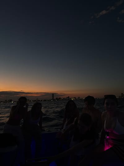 The image depicts a group of approximately seven individuals on a boat on open water at dusk, with the city of Boca del Río, Mexico, visible as a silhouette on the horizon. The sky transitions from a dark blue overhead to an orange and yellow glow at the horizon, indicative of a sunset. Sparse cloud formations are present in the upper right quadrant of the sky.
The city skyline on the horizon features numerous buildings, including several taller structures and one prominent high-rise in the center-right, with initial evening lights visible. The water surface is dark and exhibits choppy waves.
Inside the boat, which appears to be a pleasure craft, individuals are seated. Ambient blue and pink lighting illuminates parts of the boat's interior and some of the occupants.
From left to right:
1. A female subject is seated, wearing a light-colored top and bottoms. Her left arm is raised near her head, possibly holding an object or touching her ear.
2. A female subject is seated, wearing a light-colored tank top and striped pants, leaning forward.
3. A female subject is partially visible behind the second subject, obscured by shadow.
4. A male subject, shirtless, is positioned further back, facing slightly left.
5. A female subject is in the front-right, wearing a white bikini top and striped bottoms, with dark sunglasses on her head. She faces the viewer and holds a translucent object, possibly a drink container, which glows pink from an internal light source.
6. An additional individual is seated very low in the boat in the foreground, largely obscured by shadow.
7. Another individual is partially visible on the far right, also heavily shadowed.
The subjects appear to be engaged in leisure activity on the water, with subtle internal lighting visible within the boat's structure.
