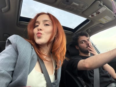 The image shows two individuals inside a car, possibly in transit, as suggested by the interior view which includes a sunroof and sun visor. The person on the left has dark hair and a beard, is wearing a black shirt, and appears to be smoking a cigarette. The person on the right has long, red hair and is wearing a white top with a gray jacket. The image suggests the setting is daytime, but the specific location within Kvemo Nichbisi, Georgia, is not discernible.