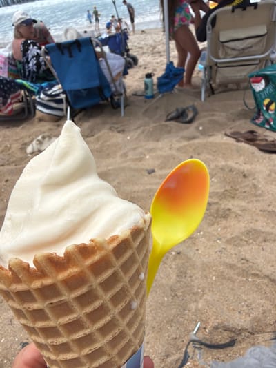 An extreme close-up shot shows a hand holding a vanilla soft-serve ice cream cone with a yellow and orange spoon positioned next to it. The ice cream is swirled in a cone with a waffle pattern. The background depicts a sandy beach with scattered beach chairs, people, and the ocean. The scene appears to be in San Clemente, California, United States of America.