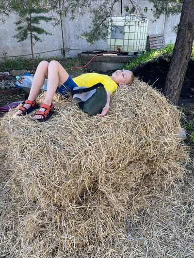 A young boy in a yellow t-shirt and blue shorts lies on his back on a large pile of hay outdoors. He wears orange sandals. A green bag is placed behind his head. The setting appears to be a rural or backyard environment in Alferyevka, Russia. In the background, there is a concrete wall, some trees, a large white water tank, and some construction materials.