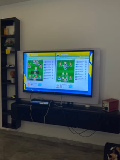 The image shows a television screen displaying a soccer team lineup. On the left side of the screen, the team "REAL MADRID" is shown with player icons and statistics. On the right side, the team "BARCELONA" is also displayed. The television is mounted above a black entertainment stand, which holds a game console and various cables. To the left of the television is a black, tall, narrow bookshelf with several shelves, containing decorative items, a toy car, and a potted plant. The scene appears to be indoors, with a light-colored wall and tiled flooring visible. The lighting is moderate, suggesting it is daytime or the room is well-lit. The overall environment seems to be a living room or entertainment area. The image does not provide any information about the city of Nabatum, Nigeria.