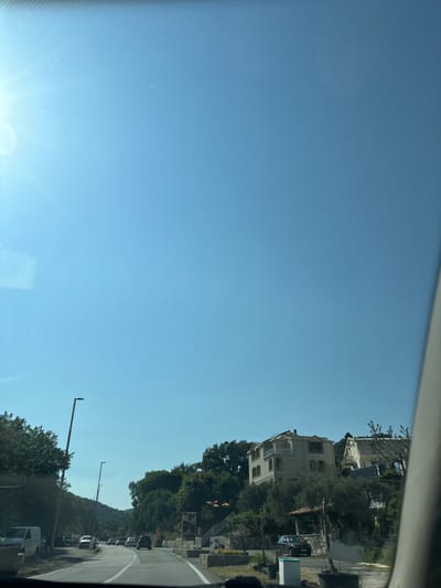 A bright sunny day in Kaluđerac, Montenegro. The image is taken from inside a vehicle, looking out through the windshield. A clear blue sky dominates the upper portion of the frame. On the right, the edge of the windshield is visible, slightly blurred. Below the sky, on a hillside, are several multi-story buildings, appearing to be houses or small apartment complexes, surrounded by lush green trees. The buildings are light-colored, with some having balconies and decorative architectural elements. A paved road winds through the scene, with several cars driving on it. There are streetlights visible along the side of the road. Trees and foliage line both sides of the road, with a van parked on the left side. The overall impression is of a scenic coastal town on a clear, warm day.