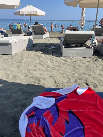 This is a ground-level, eye-level, full shot of a crowded beach on a sunny day, likely in the Germasogeia Municipality, Cyprus. In the foreground, a red, white, and purple patterned towel is draped over a dark surface, possibly a beach chair. The sand in the foreground is dark and textured, with some scattered pebbles.
In the midground, several grey lounge chairs are set up on the sand, some occupied by individuals. White umbrellas provide shade over some of the chairs. Beyond the chairs, the sea is visible, filled with numerous people swimming and wading. The water is calm and appears blue under the clear sky. In the background, a breakwater or rocky barrier extends into the sea.
Several people are visible in the water, some appear to be adults and children. A few individuals are sitting or standing on the shore. The overall impression is a lively beach scene with many people enjoying the water and sun. The presence of lounge chairs and umbrellas suggests a well-equipped beach, common in tourist areas. The lighting indicates a bright, sunny day.