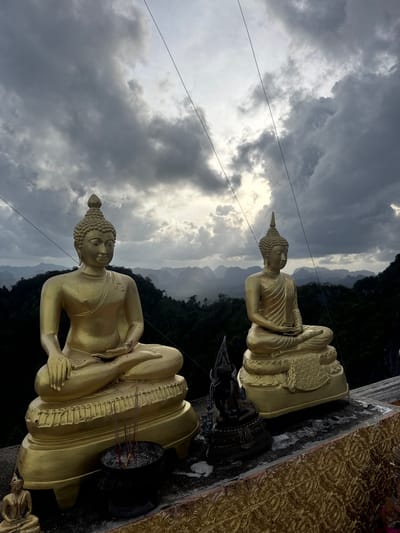 Two golden Buddha statues are positioned on a stone platform, overlooking a lush, mountainous landscape under a dramatic, cloudy sky. The statue on the left is larger and seated in a cross-legged position, with a bowl of incense sticks in front of it. The statue on the right is slightly smaller, also seated and cross-legged. In the foreground, a decorative golden railing is visible. The background is a dense forest with karst mountains in the distance, shrouded in mist. Power lines are visible stretching across the sky. The location is identified as Ban Khao Tang, Thailand.