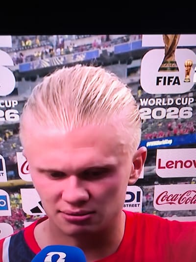 The image shows a close-up of a young man with blonde hair, wearing a red shirt. He is likely a soccer player, as evidenced by the background which features a trophy and logos related to FIFA and the World Cup 2026. The background also shows a stadium setting with spectators and various sponsor advertisements, including Coca-Cola and Lenovo. The man appears to be speaking into a microphone, suggesting he is being interviewed. The location in Kosshy, Kazakhstan, is not directly evident from the image itself.