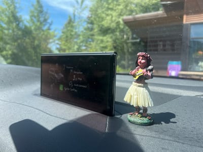 The image shows a dashboard of a car with a Hawaiian hula girl figurine to the right of the car's display screen. The screen displays menu options including "Telephone," "Navigation," "Map," "Audi connect," and "Apple CarPlay." The background, seen through the car's windshield, is a blurry depiction of trees and a building. The scene appears to be set outdoors during the day, with sunlight casting shadows on the dashboard. No specific location in Jackson, United States of America, is identifiable from the image.