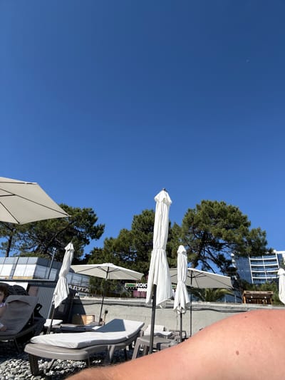 The image shows a beach scene in Adlerskiy, Russia, under a clear blue sky. In the foreground, a person's arm is visible, with part of a beach chair and the rocky shore beneath. Several white beach umbrellas are set up, some open and some closed. Behind the umbrellas, there are trees, a low wall, and buildings, including a modern hotel with a glass facade. Signs with Cyrillic text are visible on some of the buildings. A child is partially visible sitting in a lounge chair in the middle ground. The overall impression is of a sunny day at a resort beach.