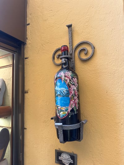 This is a close-up, eye-level, outdoor, medium shot of a decorative wine bottle hanging from a wall. The bottle is painted with a landscape scene featuring a waterfall and pink flowers. It is mounted on a wrought-iron bracket with a spiral detail. To the right of the bottle, a small sign with the words "LA LANTERNA" and "BEER WINE GARDEN" is visible. The wall is painted a textured yellow. The scene appears to be in a commercial area, possibly a restaurant or shop. The lighting suggests it is daytime, with no indication of weather.