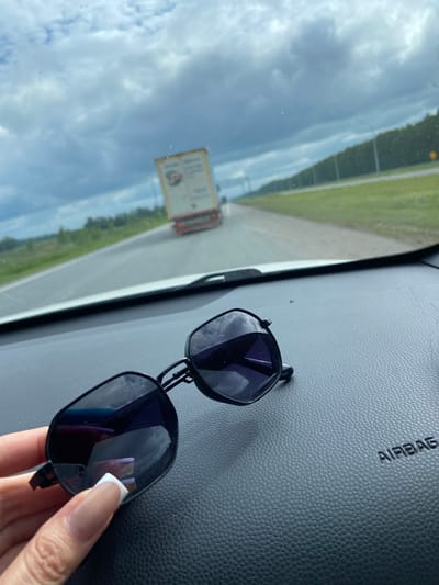An image taken from inside a vehicle shows a pair of dark-rimmed sunglasses resting on the dashboard. The sunglasses are positioned in the foreground and reflect the cloudy sky and passing landscape. In the background, a white truck with a red trailer is visible on a highway. The truck is moving away from the camera. The road is flanked by green trees and a grassy shoulder. The sky is overcast with a mix of light and dark clouds. The word "AIRBAG" is embossed on the dashboard to the right of the sunglasses. The overall impression is of a journey on a road under a moody sky. The location is identified as the city of Mirny, Russia.