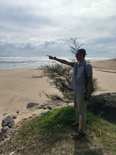 A man stands on a grassy incline overlooking a beach. He is wearing a grey polo shirt, tan pants, and black sandals. He has sunglasses on and is pointing to the left with his right arm. A bag is slung over his left shoulder. The beach is sandy and meets a choppy ocean under a cloudy sky. A thin, spiky bush is in the foreground, partially obscuring the man. Rocks and vegetation are visible on the incline.