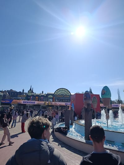 The image depicts the entrance of Europa-Park, a theme park located in Rust, Germany. A large crowd of people is entering the park. The entrance is adorned with "Bienvenue" and "Willkommen" signs and flags. Several decorative elements are visible, including a fountain with egg-shaped sculptures. Two people stand in the foreground, facing the entrance. The weather is sunny.