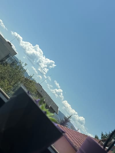 A tilted, eye-level outdoor shot captures a bright, sunny day with a clear blue sky, dotted with fluffy white clouds. In the foreground, a dark object, possibly a windowsill or frame, occupies the lower left corner, with hints of purple flowers and greenery visible. In the mid-ground, a roof with reddish-brown tiles is visible, along with evergreen trees and the side of a modern building with glass windows. Further in the background, mountains can be seen behind the trees and clouds. The scene is located in Ravno Pole, Bulgaria.