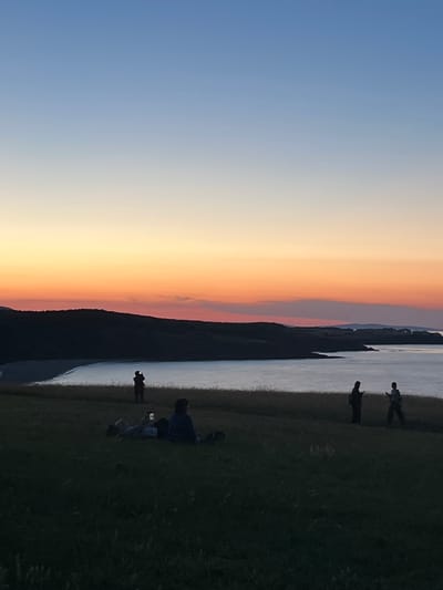 The image shows a group of people on a grassy hillside overlooking a body of water and a distant coastline. The sky is a gradient of blues and oranges, indicating sunset or sunrise.
In the foreground, on the grassy hill, several individuals are visible. To the left, two people are sitting down, one appears to be lying on their back while the other is seated. Both seem to be looking at their phones. Further to the right, two more figures are standing. One is facing left, also looking at a phone, and the other is facing right, holding something in their hands. A dark, indistinct shape, possibly a dog, is also present on the grass.
In the middle ground, on the edge of the hill where it meets the water, a single silhouette stands facing the water, possibly taking a photograph or video with a phone.
The background features a dark, hilly coastline with a calm body of water in between. The water reflects the colors of the sky. The overall scene suggests a tranquil outdoor setting, likely a park or natural area, during twilight. This scene could be depicting a location such as the area near the mouth of the Veleka River in Sinemorets, Bulgaria, known for its scenic beauty and coastal views.
