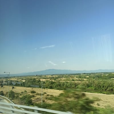 The image captures a wide-angle view of a rural landscape under a clear blue sky. The foreground features a section of a white barrier, likely from a bridge or overpass, with a blurred effect indicating movement. Two streetlights are visible on the left and right sides of the image, with the left one closer and taller. The middle ground displays a winding river or body of water, flanked by dry, golden fields and dense green vegetation. In the distance, a range of mountains forms the horizon, shrouded in a light haze. The overall scene suggests a tranquil, open environment, possibly in the vicinity of Neo Petritsi, Greece, given the characteristics of the landscape.