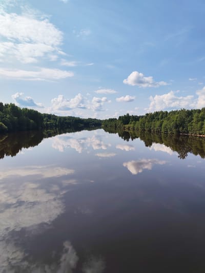 The image shows a calm river in the city of Verkhnii Rubezh, Russia, on a clear day with scattered clouds. The river's surface is still, reflecting the blue sky, white clouds, and the lush green trees lining its banks. The trees form a dense, verdant border on both sides of the river, with their reflections creating a mirror image on the water. The sky is bright blue with a few fluffy white clouds, typical of a summer day. The overall impression is one of serenity and natural beauty. There are no people or man-made structures visible in the image.