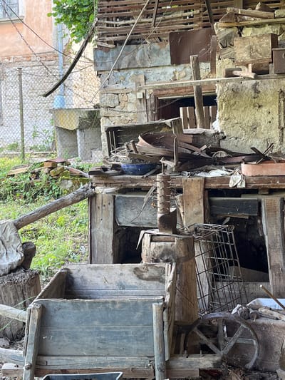 This media file captures a cluttered, semi-outdoor area, likely a storage space or a disused workshop, characteristic of a rural setting such as Byala Reka, Bulgaria.
The scene is dominated by a dense accumulation of aged materials, predominantly wood and metal, suggesting a long period of disuse or ongoing disorganization. On the right side, a large, weathered wooden structure features a prominent, rusted metal wheel with spokes, possibly from an old machine, cart, or well. Below it, a grey-blue wooden wheelbarrow or a similar large wooden container rests on the ground.
The central and left portions of the image are filled with numerous wooden planks, beams, and scraps of varying sizes and conditions, haphazardly stacked and propped against each other. Behind these, rough stone walls, possibly belonging to an old building, are visible, with some showing exposed stonework and peeling plaster. Interspersed among the wooden debris are various metal objects, including a wire mesh basket or grid, and several hanging chains.
In the mid-ground on the left, a chain-link fence runs vertically, separating the immediate area from a distant view of what appears to be part of another building with a peach-colored facade and more green foliage. A thick, dark wooden beam extends diagonally across this section. The ground level throughout the scene is covered with wild grass and weeds, indicating natural growth in an untended environment.
The natural light suggests it is daytime. There are no people visible, and no specific activity is taking place, contributing to a sense of stillness and neglect. No discernible text is visible on any of the objects. The overall impression is one of forgotten utility and rustic decay, reflecting a passage of time in an abandoned or passively maintained space.