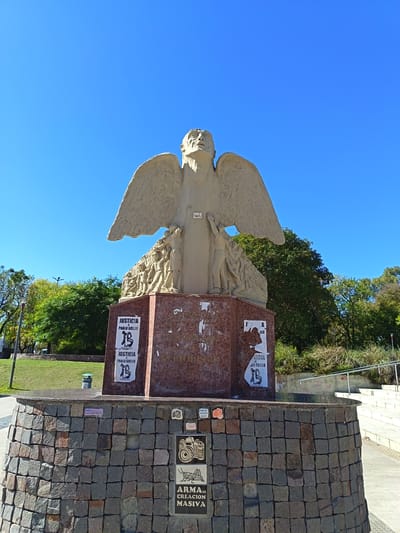 This vertical shot captures a prominent outdoor monument under a clear, bright blue sky in Remedios de Escalada, Argentina. The central subject is a large, light-colored statue resembling an angel or winged human figure, gazing upwards. Below its outstretched wings, the statue's body features bas-relief carvings of numerous smaller human figures, suggesting a narrative or collective experience.
The statue stands on a multi-tiered base. The first level is a reddish-brown marble pedestal, which is partially covered with tattered white posters. Below this, the monument rests on a substantial, curved base constructed from rectangular blocks of dark grey and brown rough-hewn stone. Embedded into this lower stone base are several decorative tiles. One tile prominently displays the text "ARMA DE CREACION MASIVA" (Weapon of Mass Creation) alongside an illustration of a vintage camera and a grasshopper. Other tiles feature symbols such as another camera and what appears to be a rose.
The posters affixed to the marble pedestal repeatedly display the phrase "JUSTICIA x PABLO GRILLO" (Justice for Pablo Grillo), accompanied by a logo that includes a stylized 'B' or an abstract symbol. The monument is situated in a green public space, with lush green trees and grass visible in the background, contrasting with the clear sky above. The bright lighting indicates it is daytime.