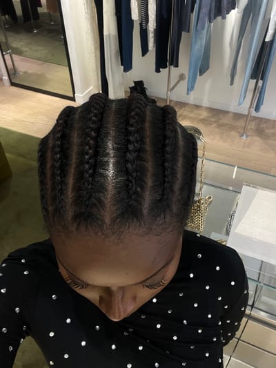An overhead image depicts an individual with dark skin, their hair styled into seven distinct cornrows running from front to back. The person is wearing a black long-sleeved garment embellished with numerous small, reflective studs. Their head is angled downwards, with the forehead, partially visible closed eyes or downward gaze, and the bridge of the nose observable.

The background indicates a retail clothing store environment located in Serris, France. Multiple metal racks are visible, displaying various garments including jeans, trousers, and tops in colors such as white, cream, dark blue, and light blue. A large, dark-framed mirror on the left reflects additional clothing and parts of the room. A pair of dark boots rests on the light-toned wooden floor near the mirror. To the right, a gold-colored, metallic link chain, consistent with a handbag strap, rests on a reflective surface, likely a glass display case. The walls are light-colored, and the scene is illuminated by interior lighting.