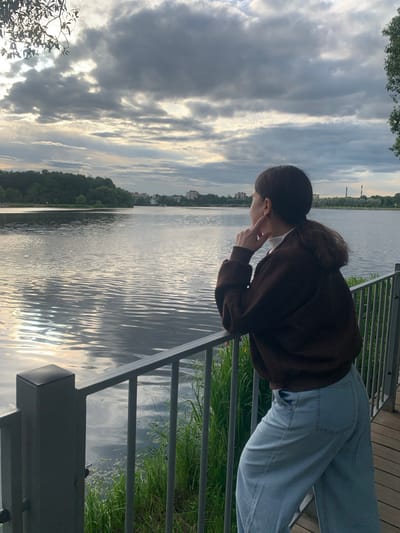 A person is leaning over a railing, looking out at a body of water. The person has dark hair pulled back and is wearing a brown hooded sweatshirt and light blue jeans. The person's face is obscured as they are looking down and slightly to the side, with one hand near their face. The scene appears to be outdoors, possibly a park or promenade, with a body of water in the background. The water has ripples, reflecting the sky, which is filled with dramatic clouds. There are trees and some buildings visible on the far bank of the water. The railing is made of dark metal, and there is wooden decking visible behind the person. The overall mood seems contemplative or perhaps curious. The lighting suggests it is late afternoon or early evening.