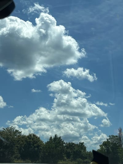 The image shows a clear blue sky with fluffy white cumulus clouds. In the foreground, a dense line of green trees forms the lower boundary of the frame. The trees appear to be deciduous and are lush with foliage. Part of a black object, possibly a graduation cap, is visible in the bottom right corner of the image. The scene suggests an outdoor setting, likely in a natural environment, given the presence of trees and the expansive sky. The location is Verinsko, Bulgaria.