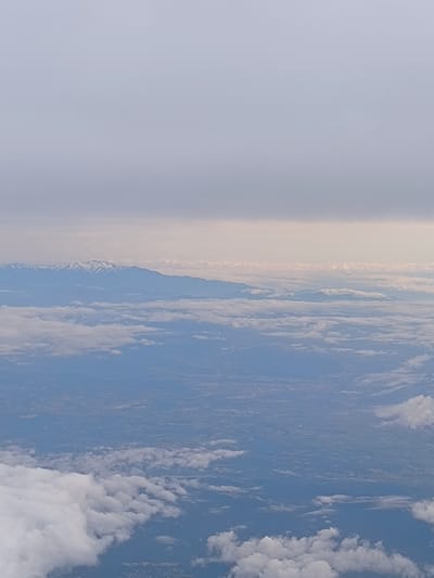 The image captures an aerial view of a landscape with distinct layers. The foreground features a collection of white clouds, varying in size and density, suggesting a high vantage point, likely from an airplane. Above this cloud layer, a discernible landmass is visible, characterized by a mosaic of fields, possible urban areas, and patches of terrain. The midground exhibits a haze or a thin cloud cover, partially obscuring the land below, giving a sense of distance.

In the background, a mountain range is prominently displayed, with some peaks covered in snow, providing a stark contrast to the surrounding hazy atmosphere. The sky above the mountains is overcast, with a uniform, grayish hue.

The location, inferred from the request, would suggest this image shows a panorama of the landscape in and around Warngau, Germany, likely with the Bavarian Alps in the background. The specific details on people, subjects, objects, actions, and interactions are limited due to the nature of the aerial perspective.