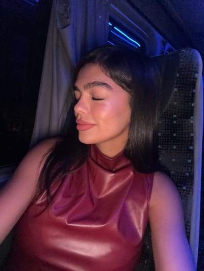 A young woman with long dark hair is sitting in a vehicle, wearing a burgundy leather top. The woman is looking down with her eyes closed, likely in a relaxed or sleepy state. The interior of the vehicle is dimly lit with a blue glow emanating from a strip of light in the background. The seat the woman is on is a dark blue with a patterned fabric. The coordinates 31.937, 34.857 locate the city of Ramla, Israel. However, the image does not provide any specific location information within the city.