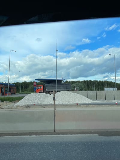 The image depicts a scene in Bykovka, Russia, showing a roadside view. A gray construction truck is visible. A pile of gravel is along a concrete barrier next to an asphalt road. The sky is blue with scattered white clouds. Utility poles are present. A Drive Cafe is also visible in the lower part of the image. A stop sign is next to the construction truck. A forested area is in the background.